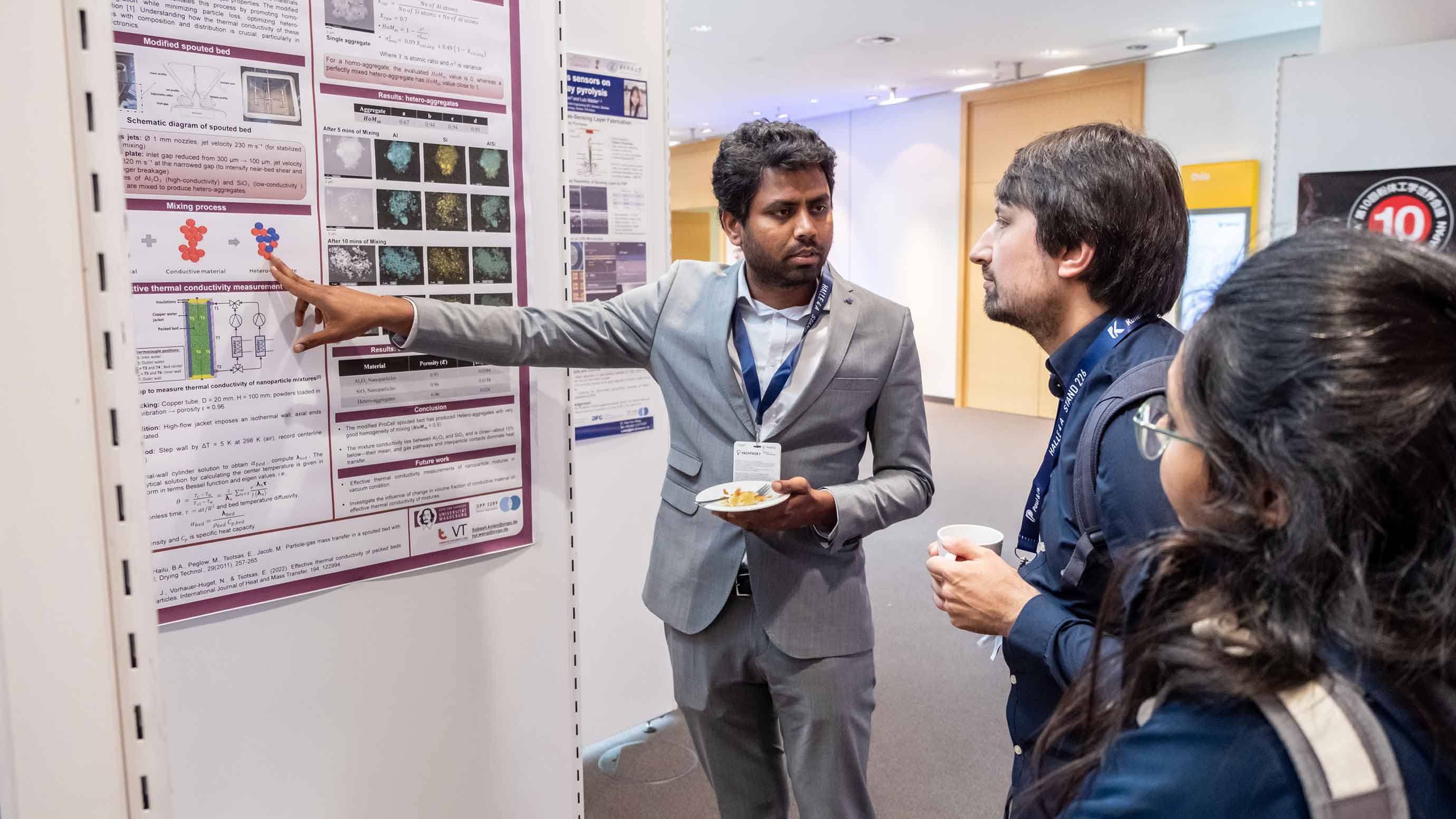 A person stands beside a scientific poster, pointing to a section of the displayed research while speaking with two other attendees. All three individuals wear conference badges, and one person holds a cup while another holds a small plate. The scene takes place in a brightly lit exhibition or conference hallway with additional posters visible in the background.