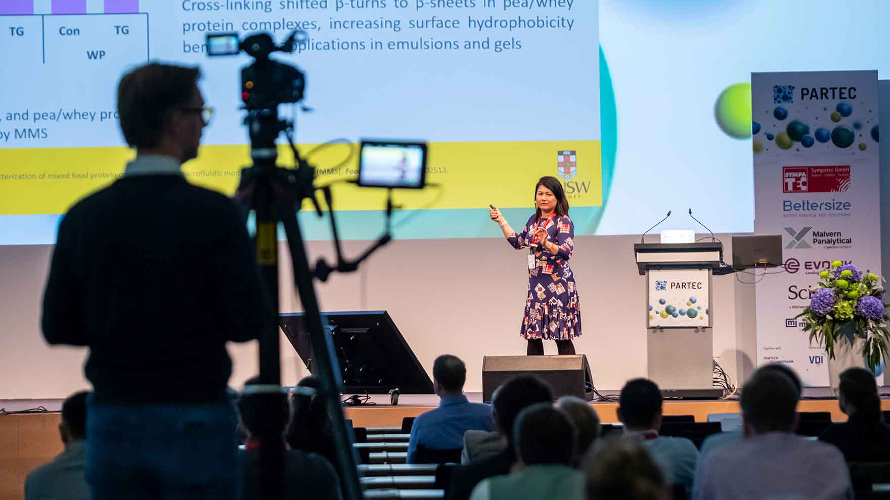 A person stands on a conference stage giving a presentation in front of a large projected slide containing scientific text and diagrams. Audience members sit facing the stage, while a camera operator records the session from the side. A podium labeled “PARTEC” and a display stand featuring various sponsor logos are positioned on the right.