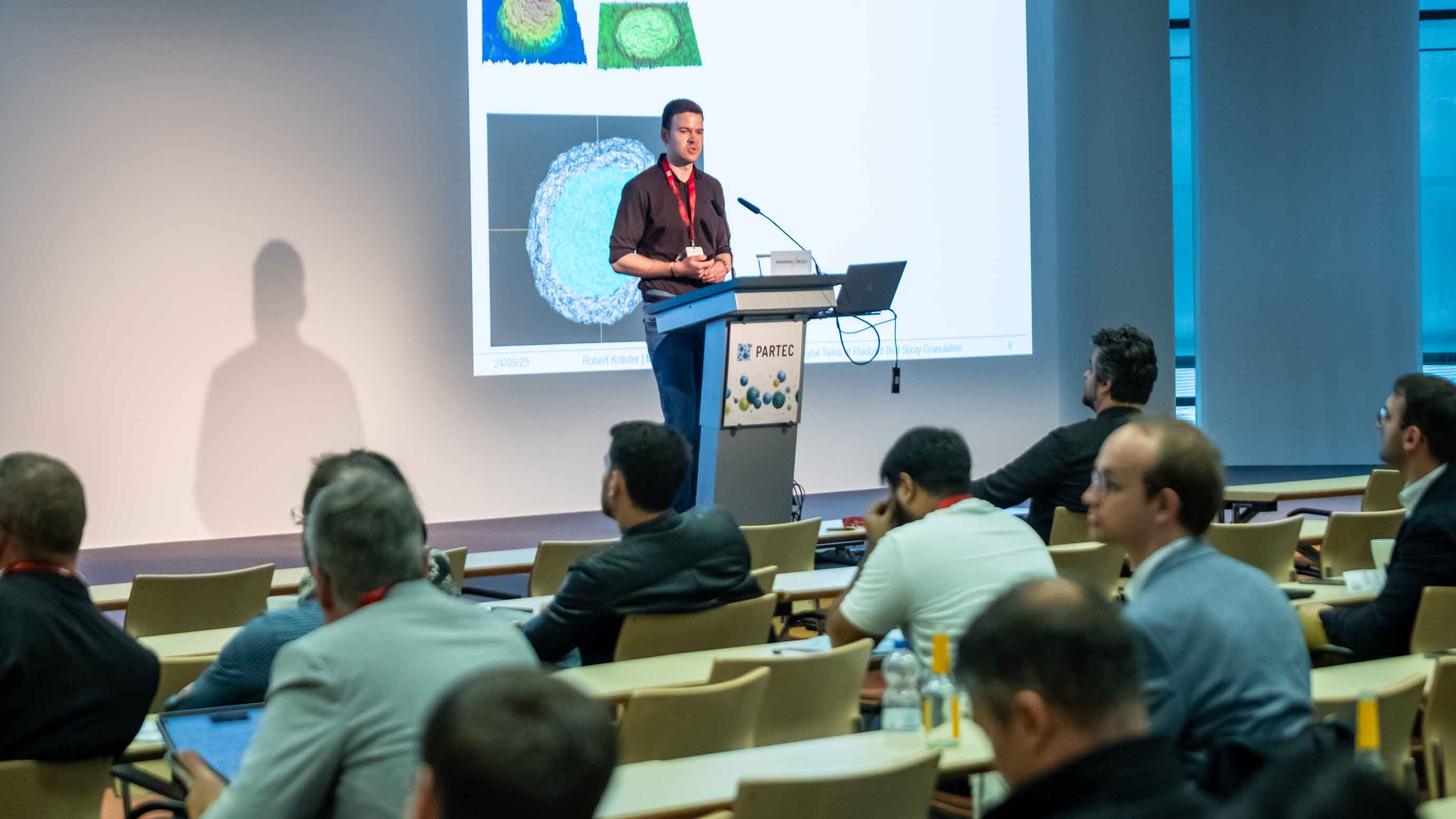 A person stands at a conference podium labeled “PARTEC,” giving a presentation in front of a projected slide showing scientific imagery. Several attendees sit in rows facing the stage, and a camera is positioned on the right side recording the session.