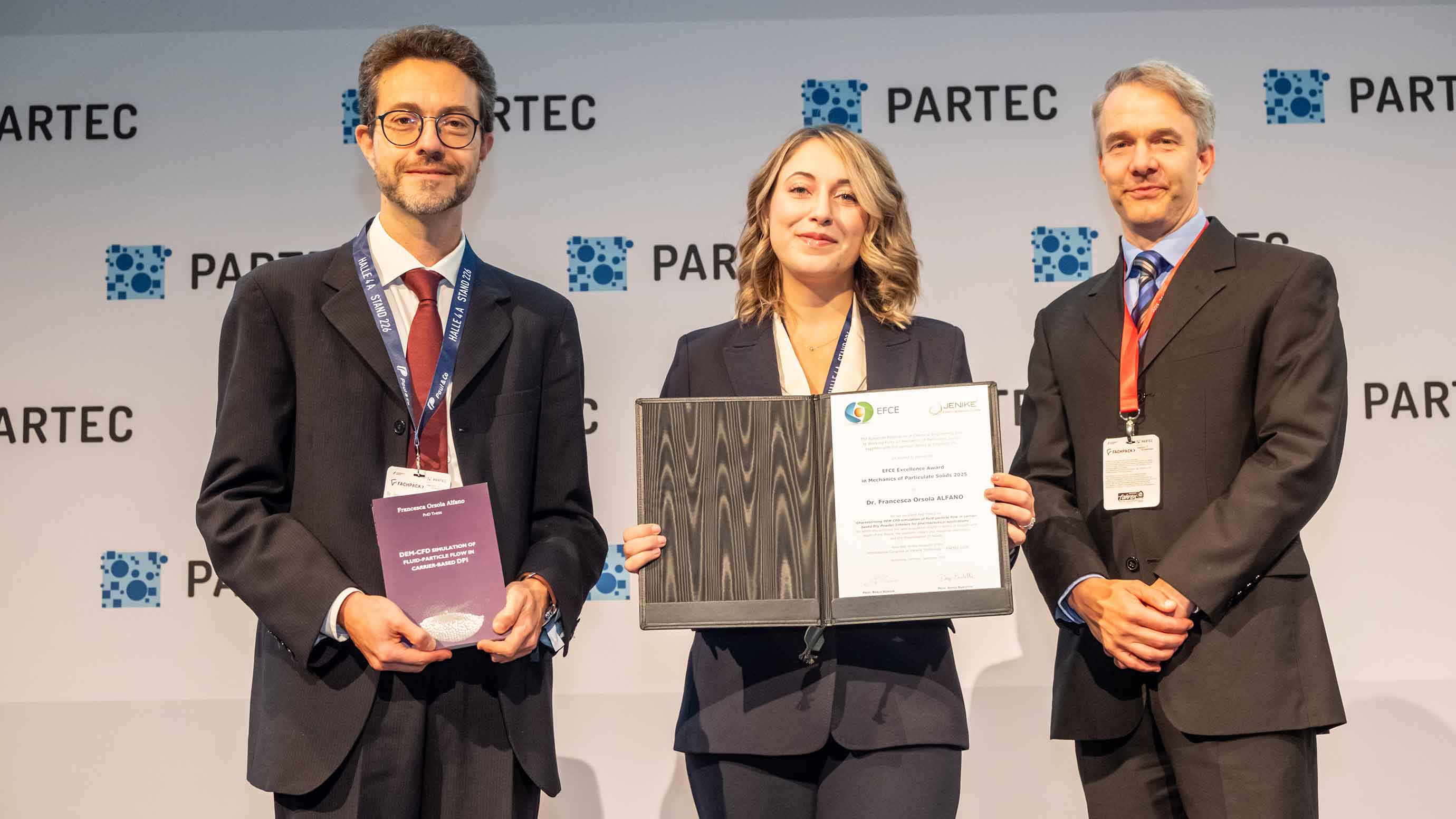 Three people at PARTEC event, one holding an award, standing in front of branded backdrop.